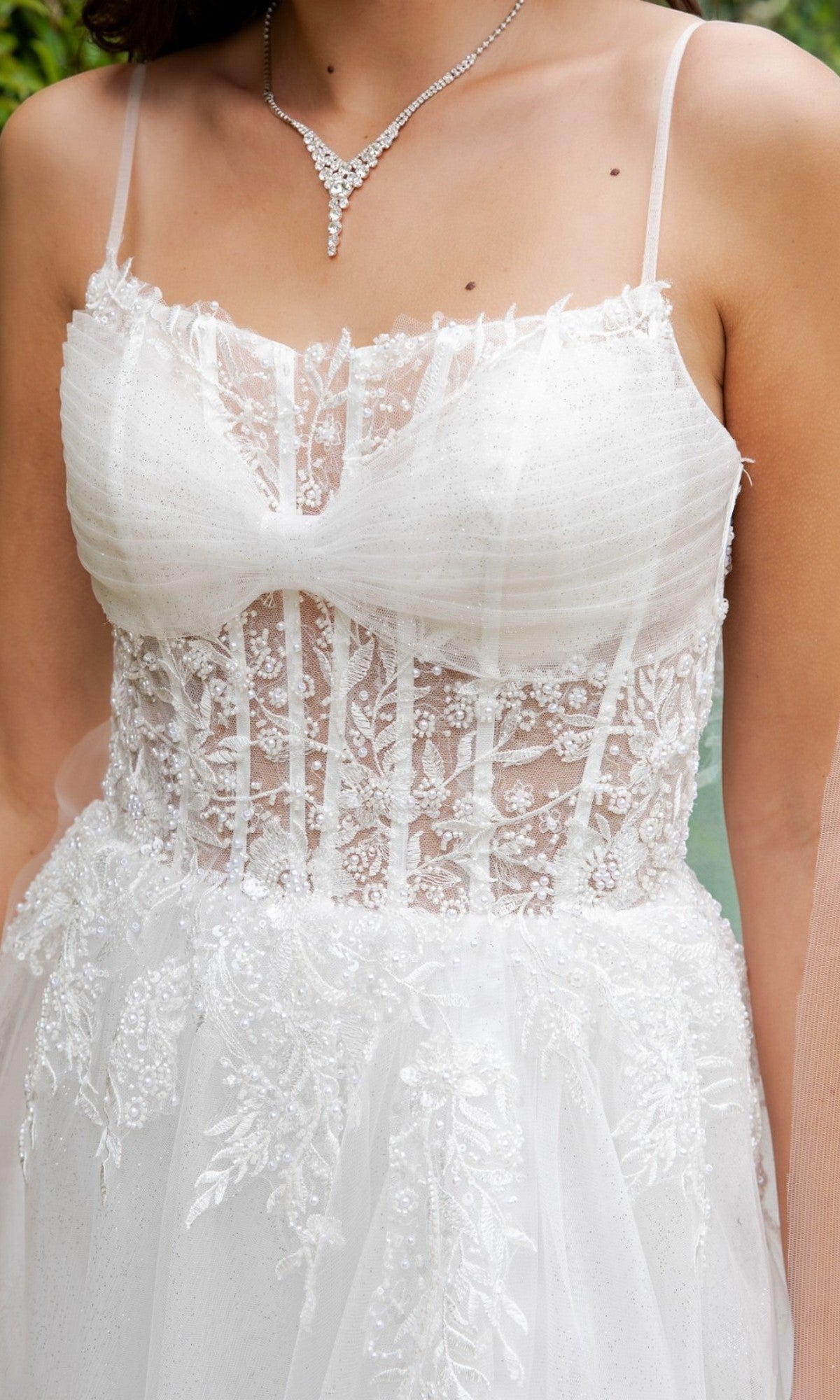 A woman wearing the Cinderella Couture 4217, a long formal white bridal gown with thin straps, crafted from sheer lace adorned with a floral pattern and delicate pearl-beaded details, paired with a silver necklace.